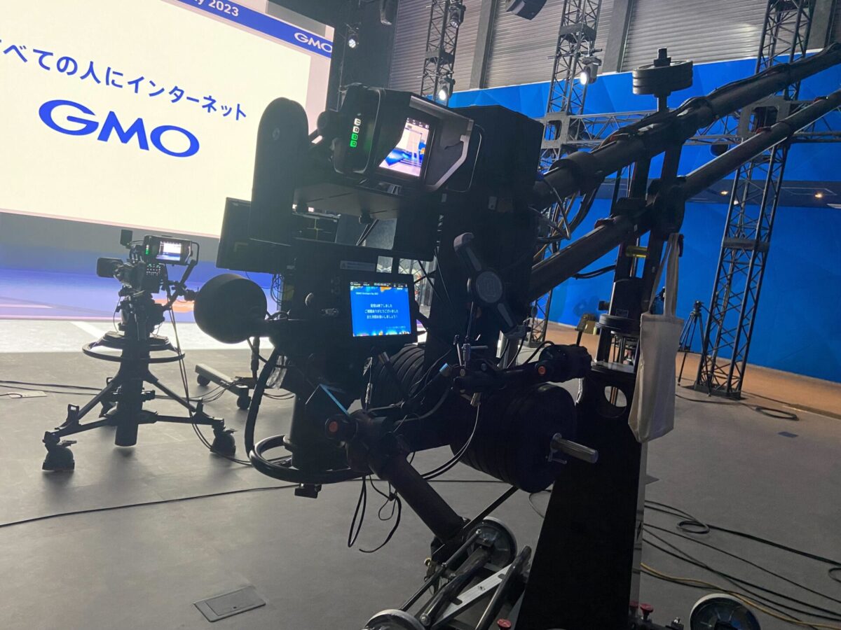 GMOグローバルスタジオに設置された大型カメラジブ(クレーン)システム。手前にはカメラのパン・チルトやズームを操作するモニターとコントローラーが配置され、奥にはもう一台のカメラと「GMO」のロゴが表示された背景スクリーンが見える。プロフェッショナルな配信・収録環境の様子。